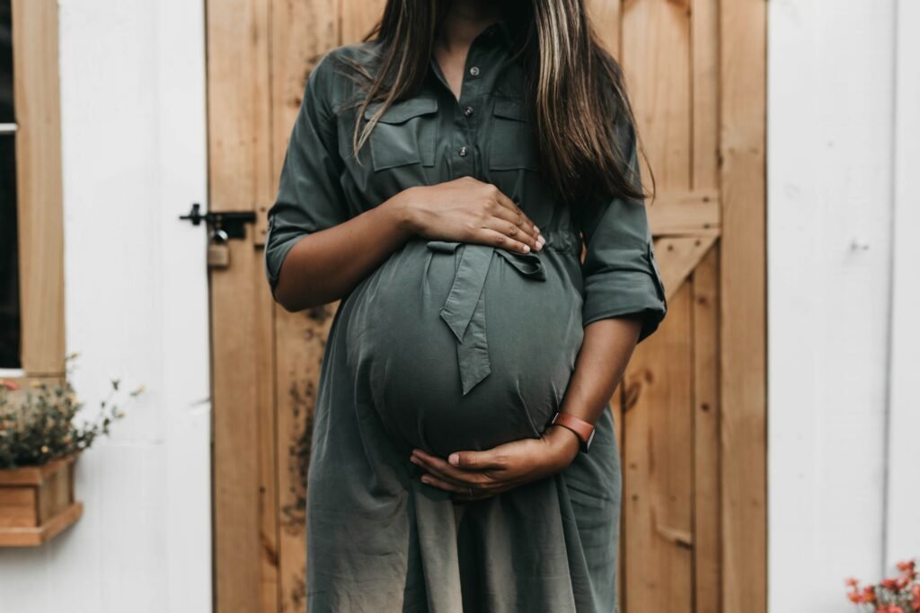 Pregnant woman in a soft olive-green dress gently cradling their belly, set against a backdrop of sunlit green leaves.